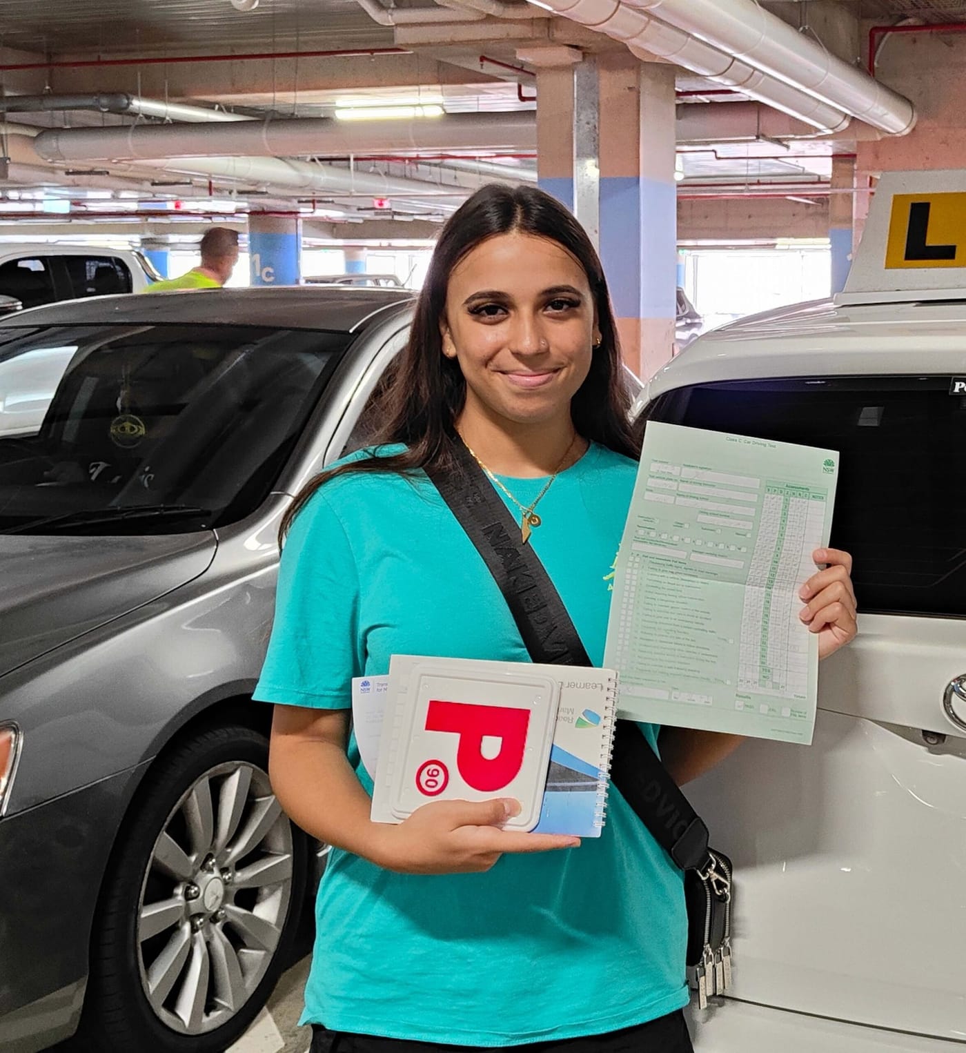 A new driver holding P plates and paperwork in a car park beside a learner car